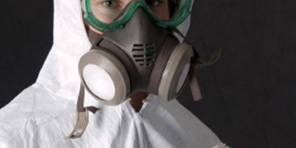 A person wearing a protective suit, green goggles, and a respirator mask appears to be conducting an investigation. In a setting requiring safety gear, one blue-gloved hand holds a piece of cloth. The background is dark and foreboding.