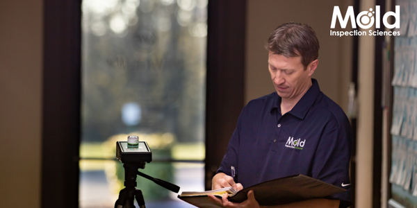 A person in a navy blue shirt with the Mold Inspection Sciences logo is writing on a clipboard, deep in investigation. Next to them is a monitoring device on a tripod. In the background, there's a blurred window with sunlight filtering through.