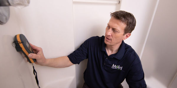 A person in a navy shirt uses a handheld moisture meter on a white shower wall, testing for mold and checking for dampness.