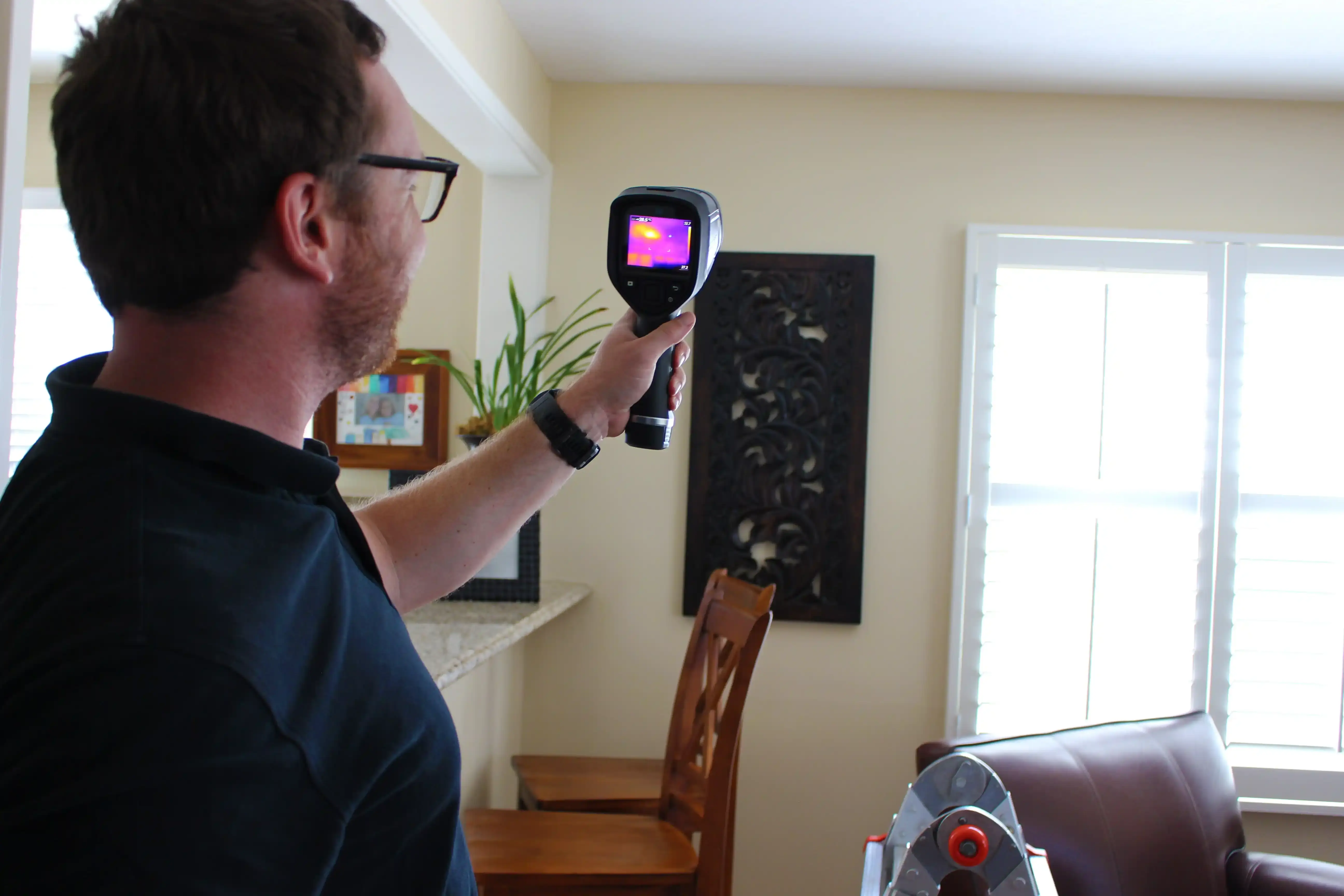 A person wearing glasses holds a thermal imaging camera in a living room, actively testing for heat variations. The camera screen displays a vibrant heat map. In the background, there are chairs, a window with blinds, and wall art.