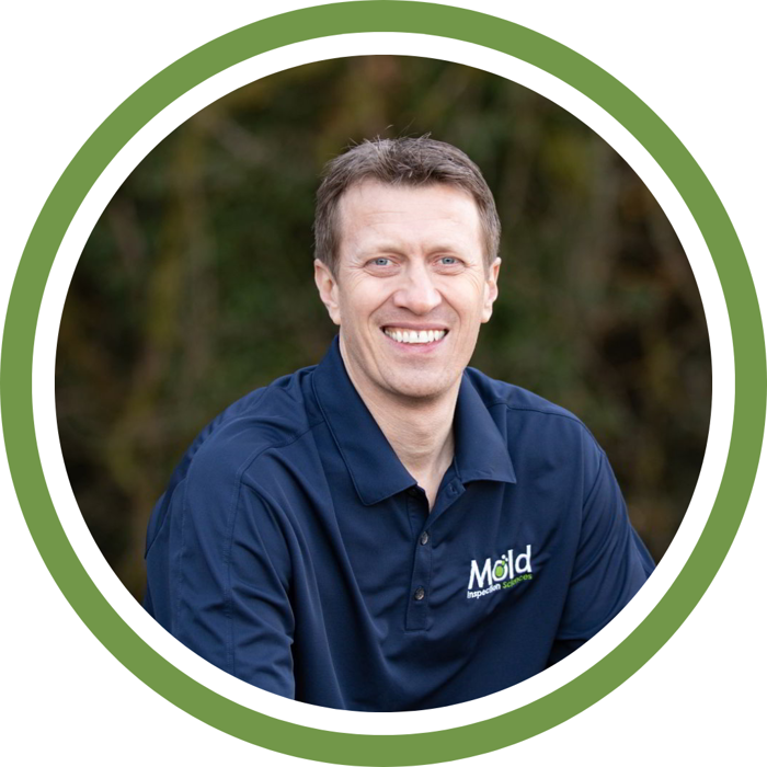 A person with short hair is smiling while wearing a dark blue polo shirt that features Mold and a leaf design, reminiscent of nature's assessment. The image is framed by a circular green border, while the blurred background hints at greenery.