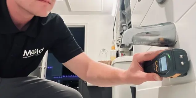 A person in a black polo shirt with a Mold Inspection logo conducts an inspection, using a moisture meter on white bathroom tiles near a bathtub faucet.