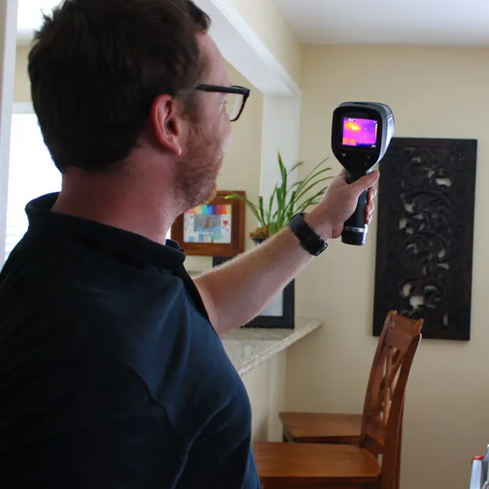 A man with glasses conducts an inspection using a handheld thermal imaging camera inside a room. The screen reveals a heat signature. He's wearing a dark shirt, and in the background, wooden chairs and a decorative wall hanging can be seen.
