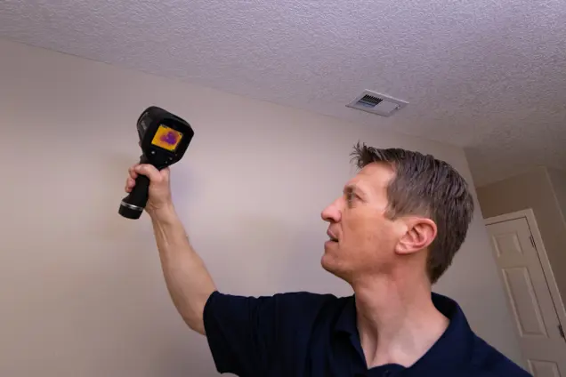 A person holding a thermal imaging camera is conducting an inspection of a wall near a ceiling vent. The screen reveals varied temperature zones, suggesting potential mold presence. With focused attention, the investigator examines the light-colored room with its closed door.