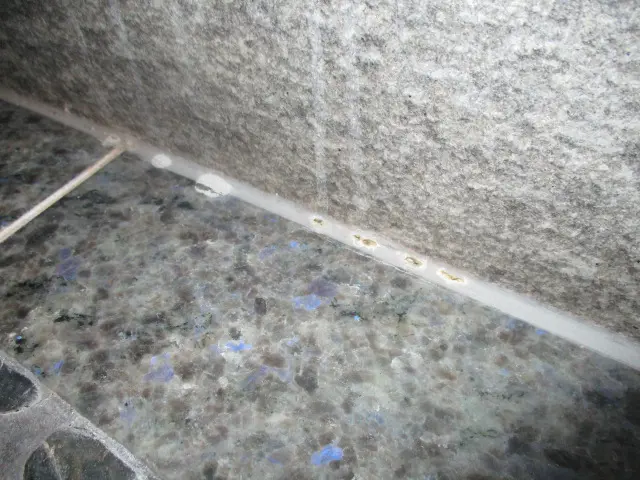 Close-up of a stone wall and a polished stone countertop with a visible seam. Upon inspection, the seam is sealed with a strip of white caulking, which shows slight discoloration. There are small yellow stains along the caulking line, possibly indicating mold growth.