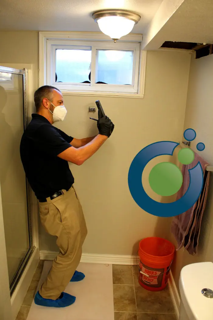 A person wearing a mask, gloves, and shoe covers is carefully photographing a bathroom corner for mold testing. There is a small window above and a bucket on the floor. A logo with overlapping circles is partially visible on the right wall.