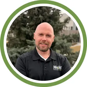A smiling bald man with a beard, wearing a black Mold Inspection Sciences polo shirt, stands outdoors in front of a blurred evergreen tree. The image, framed by a green and white circular border, highlights his expertise in inspection and investigation.