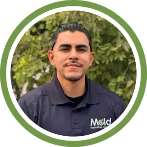 A man with dark hair and a trimmed beard, involved in mold inspection and testing, wearing a navy polo shirt with a Mold Inspection Sciences logo, stands outdoors in front of leafy green plants. The image has a green circular border.