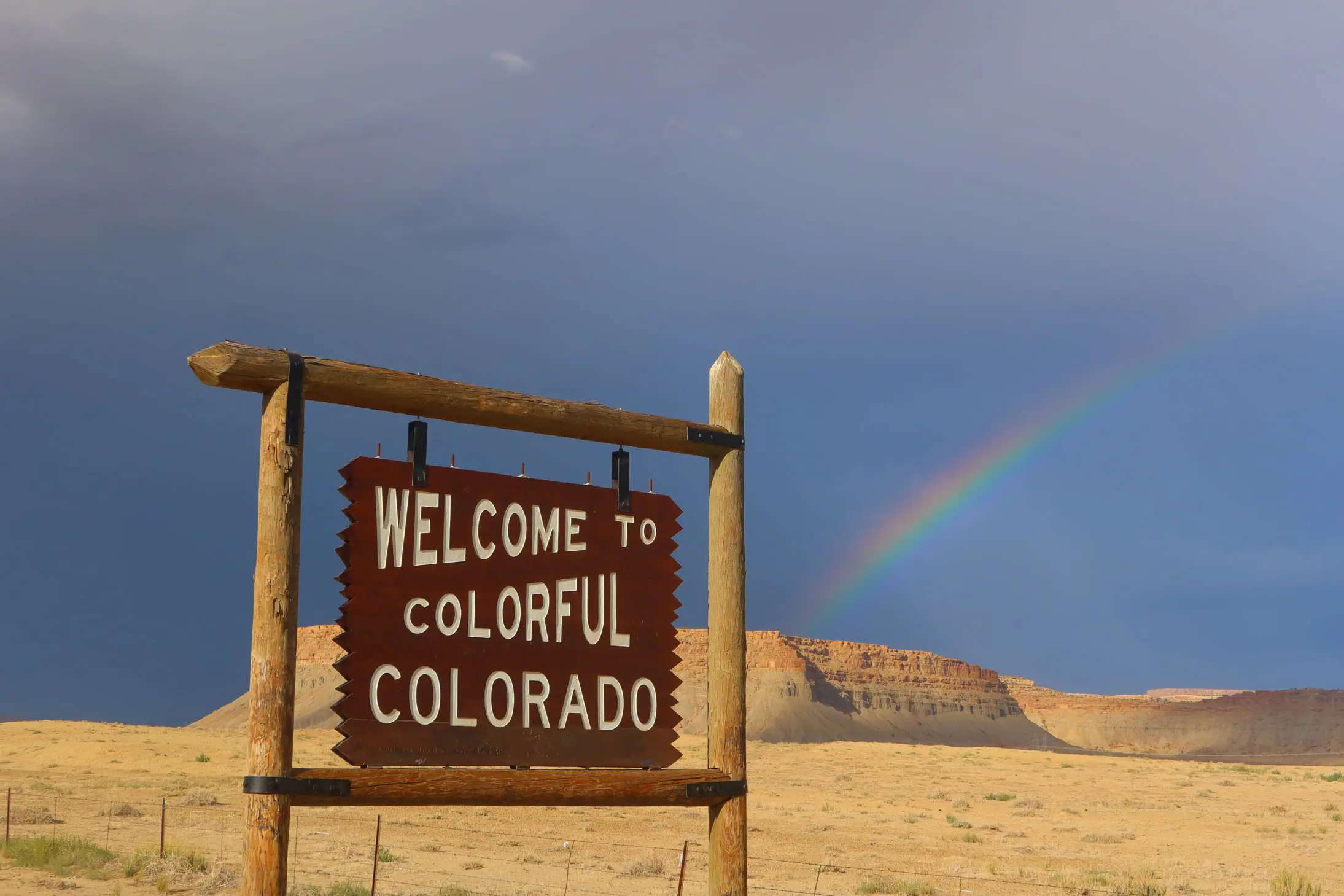 A wooden sign reads "Welcome to Colorful Colorado" against a backdrop of a desert landscape with rocky cliffs, as if nature's meticulous sampling. A vibrant rainbow arches across the deep blue sky.