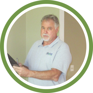 A middle-aged man with gray hair and a beard stands indoors, clipboard and pen in hand, ready for a mold inspection. He wears a light gray polo shirt with an embroidered logo framed within a green circle border, exuding professionalism.