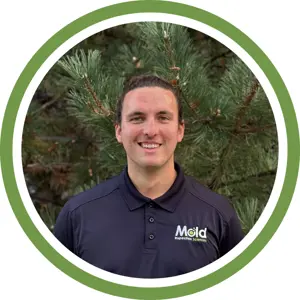 Smiling person with brown hair in a ponytail, wearing a dark polo shirt with Mold Inspection Sciences logo, stands in front of green pine tree branches, representing expertise in inspection within a green and white circular frame.