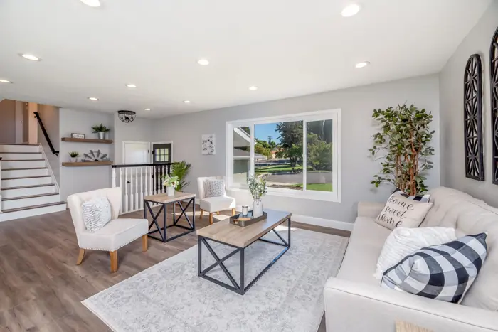 Spacious, bright living room with wooden flooring, beige sofa, two armchairs, and a wooden coffee table. Large window overlooks a green yard. Stairs lead to the upper level. Decor includes plants and wall art—perfect for a relaxing retreat or an informal inspection of comfort and style.