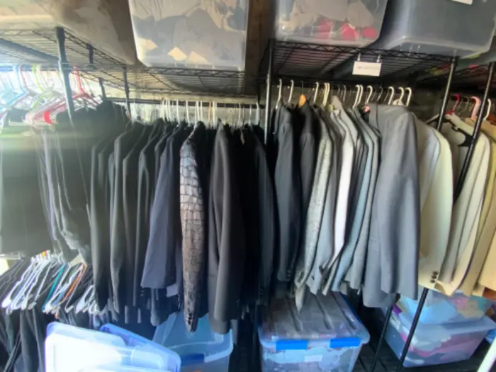 A closet with wire shelving holds neatly hung suits in various shades of black, gray, and light colors. Clear plastic bins, recently passed inspection for mold, are stored above and below the racks.