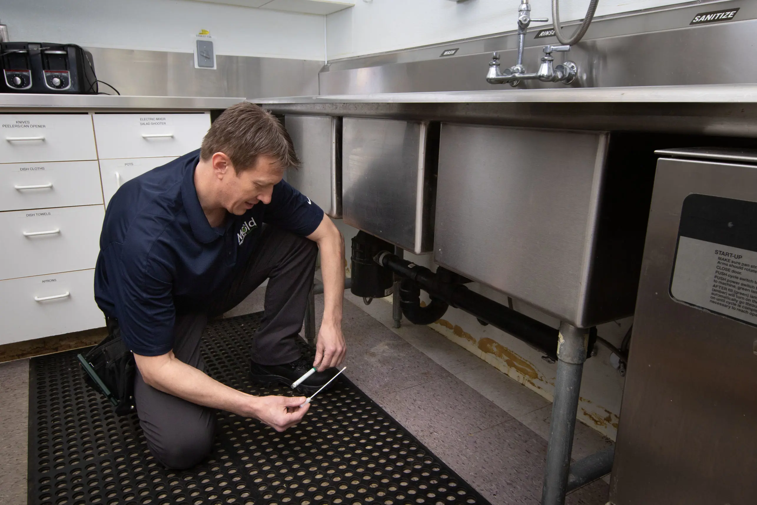 A man crouches beside a stainless steel sink, adjusting pipes with a wrench. The setting, possibly undergoing mold assessment, appears to be a commercial kitchen or utility area, featuring rubber mat flooring and cabinets.