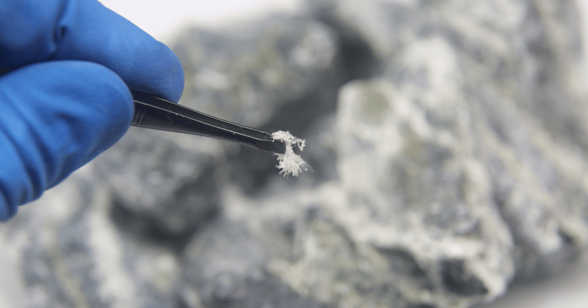 A gloved hand holding tweezers grips a small, fibrous mineral sample against a blurred background of grayish rocks, meticulously poised as if conducting an investigation.