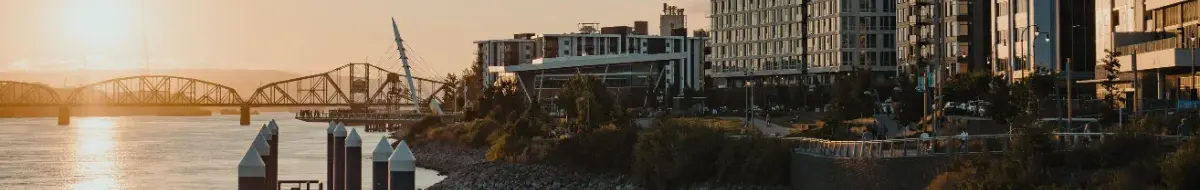 Sunset view of a waterfront cityscape with modern buildings, a bridge over a river, and a paved walkway lined with greenery—ideal for mold sampling or environmental testing as sunlight reflects off the clear water.