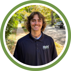 A person with long hair and a mustache smiles outdoors, embodying the spirit of inspection. They wear a black polo shirt with the Mold Inspection Science logo. The background features trees and a sidewalk under a bright sky, framed by a green circle.