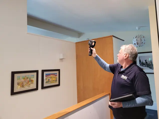 A man in a dark polo shirt uses a handheld infrared camera, pointing it towards a ceiling corner in the room for an investigation. He holds a tablet in the other hand. The room, with light-colored walls and framed artworks, is being assessed for potential mold issues.