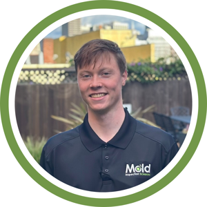 A smiling person is standing outdoors, wearing a black polo shirt emblazoned with the company logo. Behind them, a wooden fence and urban buildings set the scene for their latest mold inspection endeavor.