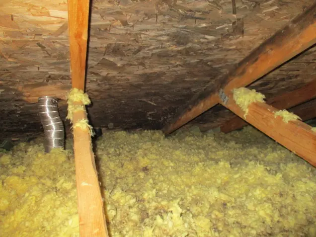 An attic with wooden beams and yellow insulation covering the floor, ideal for sampling. A ventilation duct extends from the floor to the ceiling, which is made of chipboard panels.