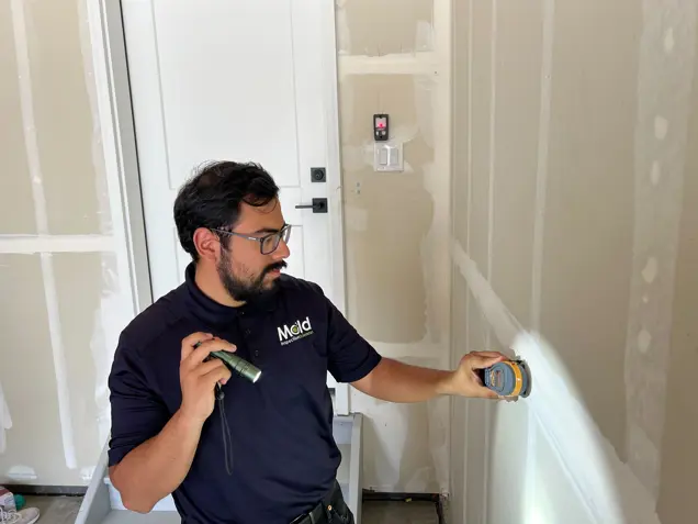 A man wearing glasses and a black Mold shirt uses a flashlight and a handheld device to conduct an inspection of drywall in a room under construction or renovation.