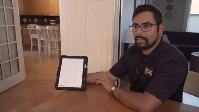 A man with glasses sits at a wooden table, holding up a tablet displaying a mold investigation list. He wears a black collared shirt with a logo, in a home setting with the kitchen visible in the background.