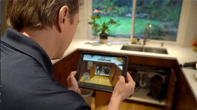A person holds a tablet displaying a virtual overlay for inspection of supply lines in a kitchen under-sink cabinet. The kitchen features wooden cabinets, a silver faucet, and a window with a view of greenery.