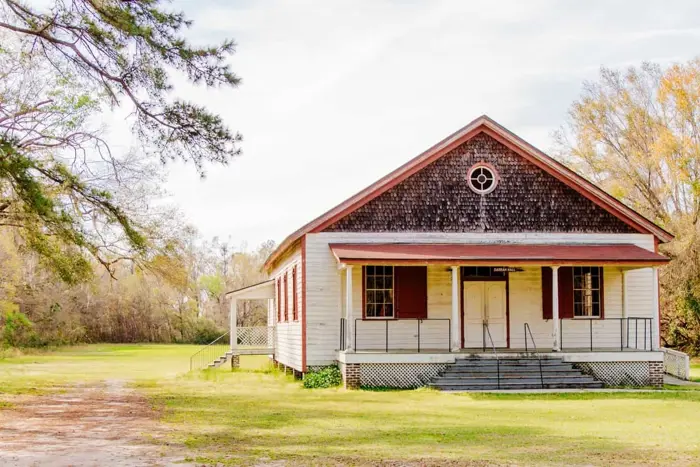 A rustic, wooden building with a triangular roof and red shutters sits amidst a grassy field. Surrounded by trees, the structure offers a front porch with steps leading up to double doors. An ideal setting for sampling serenity under the round window adorning the gable.