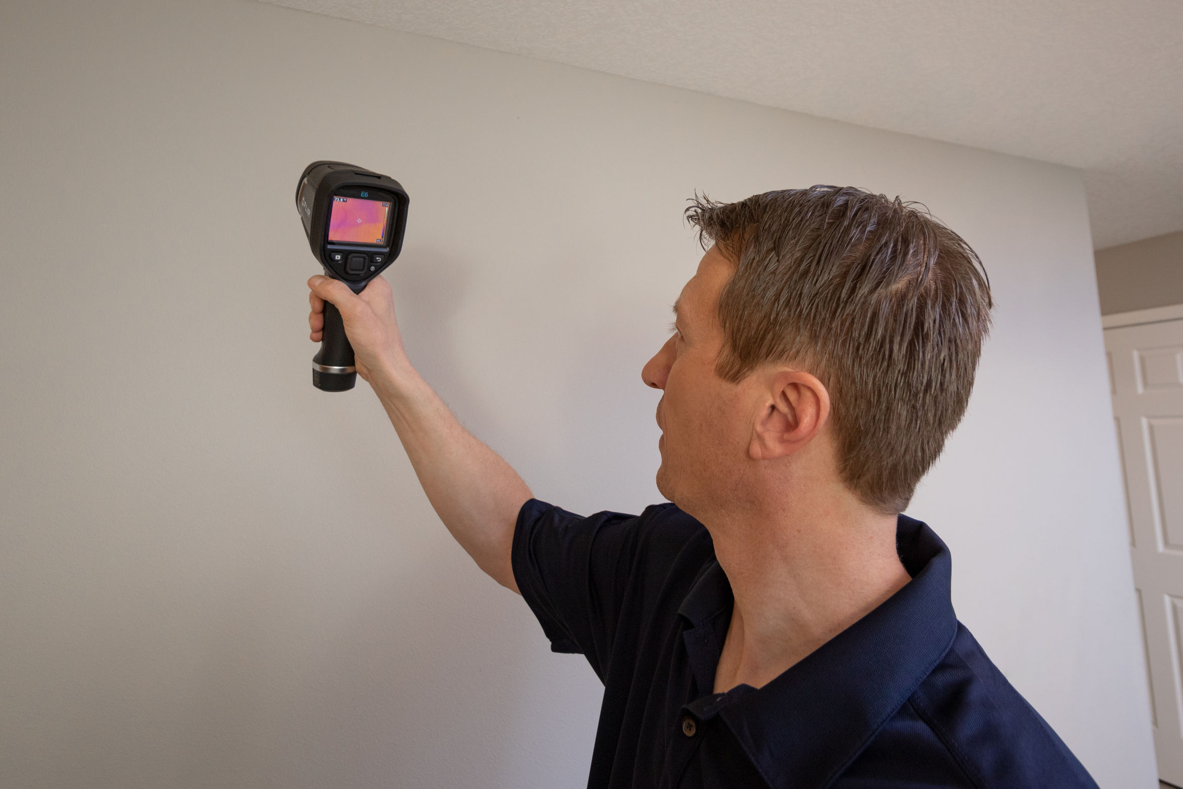 A person conducts an inspection using a handheld thermal camera to scan a white wall in the room. The camera's screen reveals a vivid heat map, ranging from red to blue. Wearing a dark shirt, the individual is intently focused on assessing the wall's temperature variations.