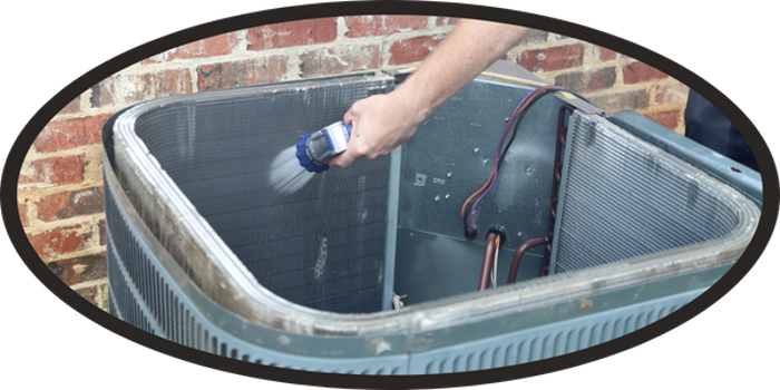 A person sprays water to clean the inside coil of an outdoor air conditioning unit next to a brick wall as part of regular assessment and maintenance.