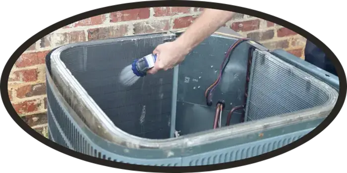 A person sprays water to clean the inside coil of an outdoor air conditioning unit next to a brick wall as part of regular assessment and maintenance.