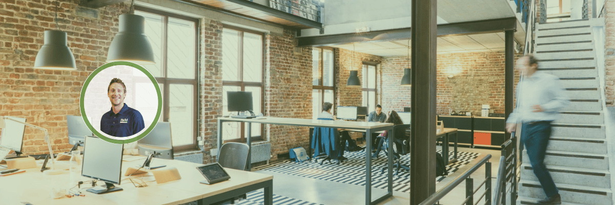 A modern office space with exposed brick walls, several desks with computers, and a striped rug evokes a lively vibe. Three people are working, perhaps conducting an assessment, as one person walks up the stairs. An inset image captures someone smiling against a bright background.