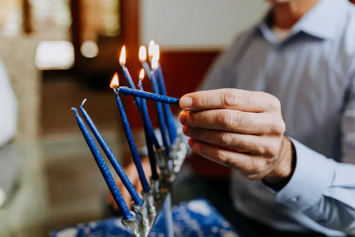 A person is lighting blue candles on a menorah with a white candle, as if conducting an assessment of tradition. The scene suggests a Hanukkah celebration in a warmly lit room, with some candles already burning. The menorah stands proudly on the table.