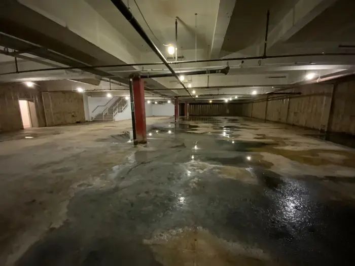 A dimly lit, empty underground parking garage with a wet concrete floor hints at its worn and unused state. The industrial-style ceiling and support columns stand in silent inspection, while the staircase and open doorway suggest a space awaiting new purpose.