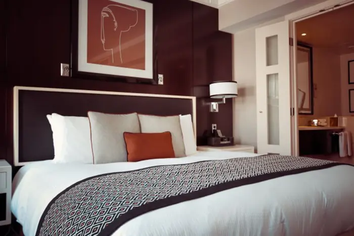 A modern hotel room featuring a neatly made bed with white linens and decorative pillows. A patterned blanket is folded at the foot. The room includes dark wood accents, wall art, and a view into a bathroom with open doors, meticulously tested to ensure a pristine stay.