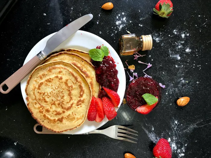 A plate of pancakes with strawberry slices, raspberry jam, and mint leaves on a black countertop. A small bottle of syrup, a knife, a fork, almonds, and scattered flour are around the plate, creating the perfect setting for testing your breakfast sampling skills.