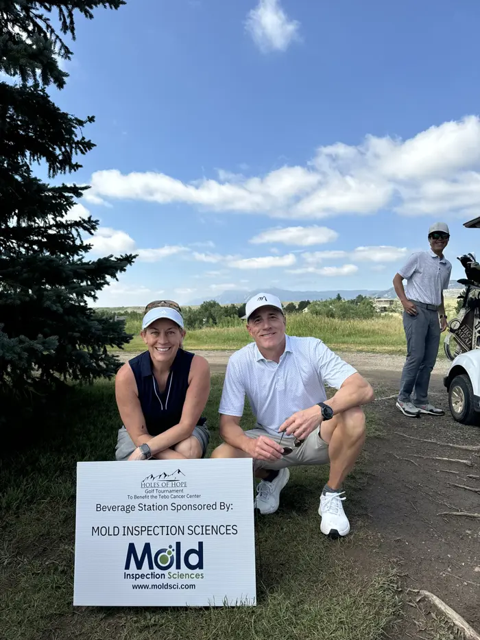 Two smiling people in athletic wear crouch by a sign for Mold Inspection Sciences, indicating sponsorship of a beverage station. They're on a lush grassy area with a golf cart and another person in the background under a partly cloudy sky, showcasing their commitment to detailed inspection.