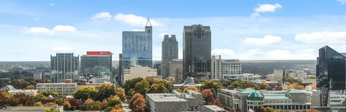 A skyline view of a city with tall buildings under a blue sky and scattered clouds. The foreground, resembling an artist's assessment of nature, shows colorful autumn trees and smaller structures, creating a vibrant contrast against the modern skyscrapers.