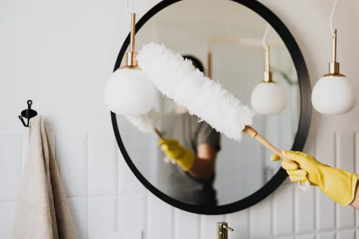 A person wearing yellow gloves uses a fluffy duster to meticulously inspect a round mirror. A beige towel hangs nearby on a hook, and two round pendant lights add ambiance. The setting appears to be a bathroom, meticulously prepared for investigation or sampling activities.