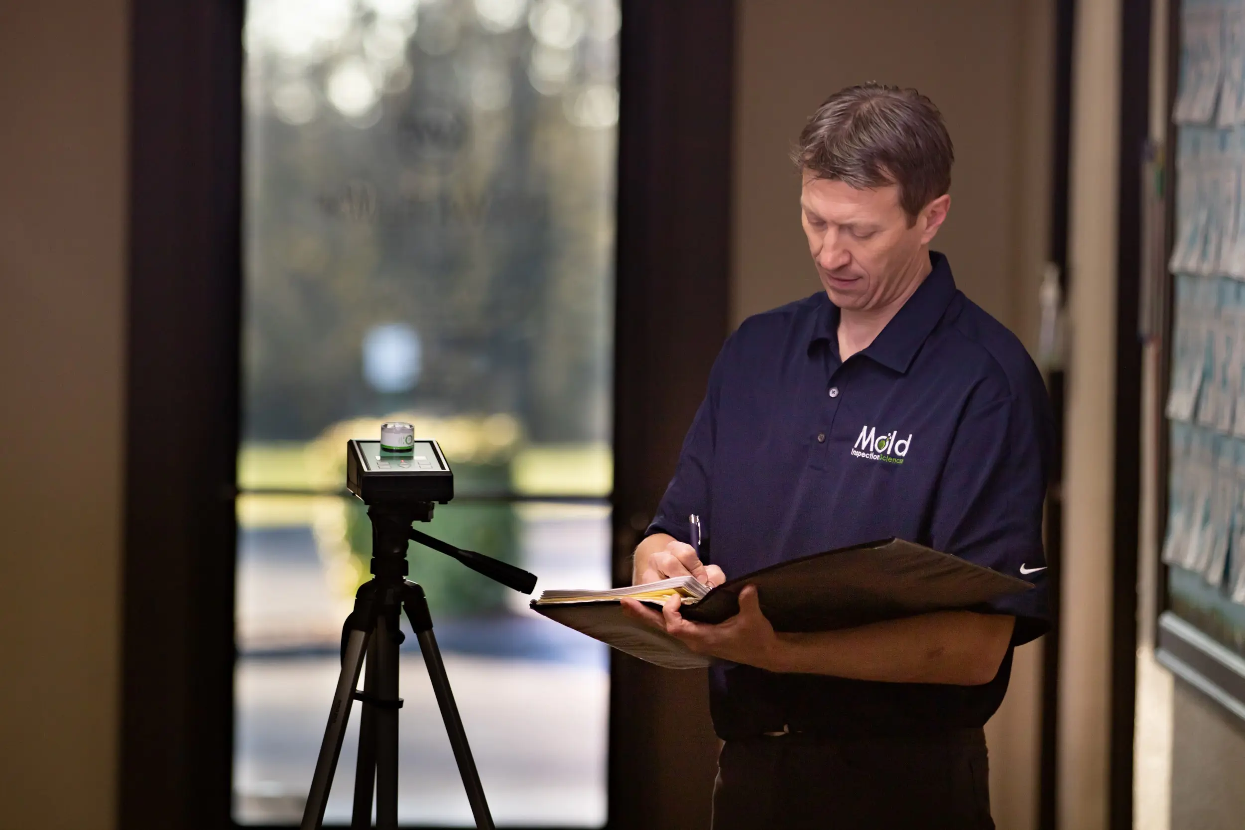A man in a navy blue shirt is standing indoors, sampling data as he writes on a clipboard. A camera on a tripod is positioned in front of him, possibly recording the testing process. The blurred background shows a door leading outside with sunlight streaming in.