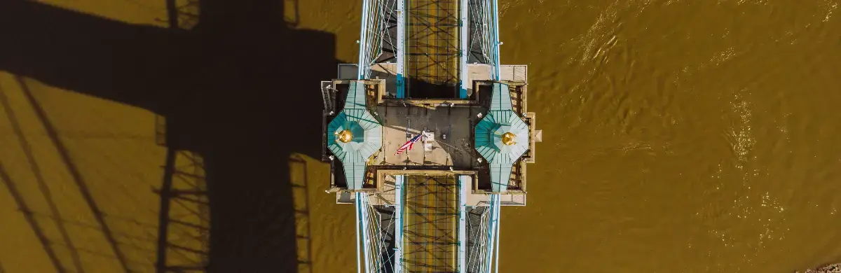 Aerial view of a suspension bridge with a distinctive blue and teal design, casting a large shadow over a brownish river. An investigation flag flutters in the middle of the bridge.
