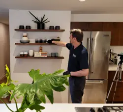 A man in a dark polo shirt carefully arranges items on a wooden shelf in a modern kitchen, conducting an assessment of the space. The shelves hold plants and various small objects, while a large green plant stands prominently in the foreground, framed by a stainless steel refrigerator behind.