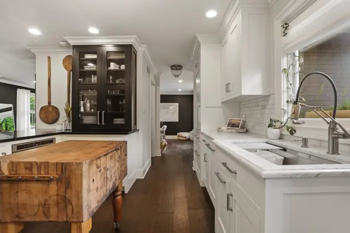 Modern kitchen with white cabinetry, marble countertops, and stainless steel fixtures. A wooden butcher block island sits in the center, perfect for sampling culinary creations. A glass cabinet displays dishes while a large window provides natural light. Dark wood flooring completes the look.