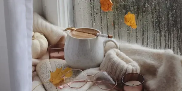 A cozy scene by a rainy window with a mug of coffee, an open book, glasses, a candle, a sweater, and autumn leaves scattered around—perfect for reading or jotting down notes about mold testing and sampling on a crisp autumn day.