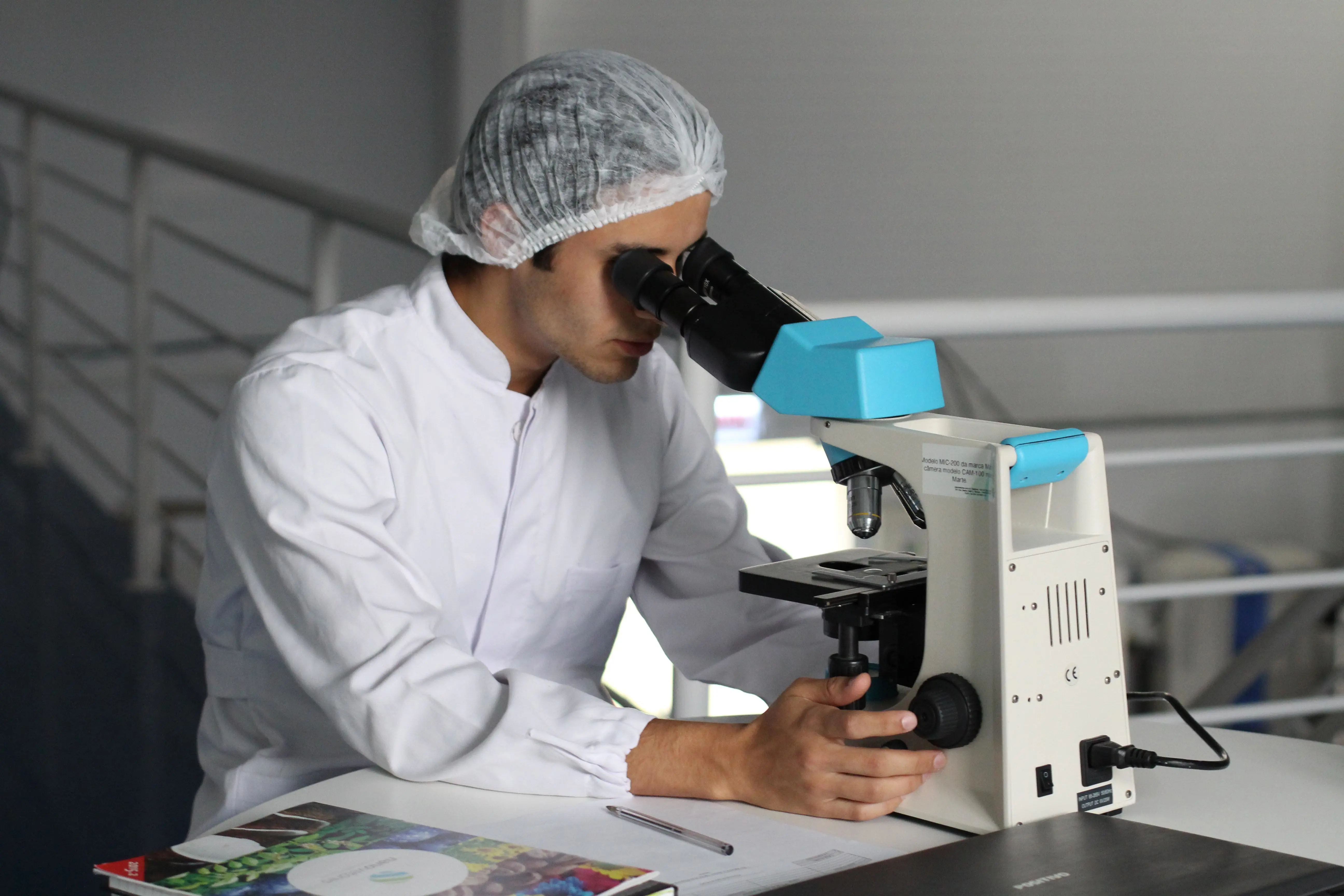 In a laboratory setting, a person in a white lab coat and hairnet meticulously conducts an investigation through a microscope. Nearby, a pen and document suggest detailed findings on mold contamination are being documented.