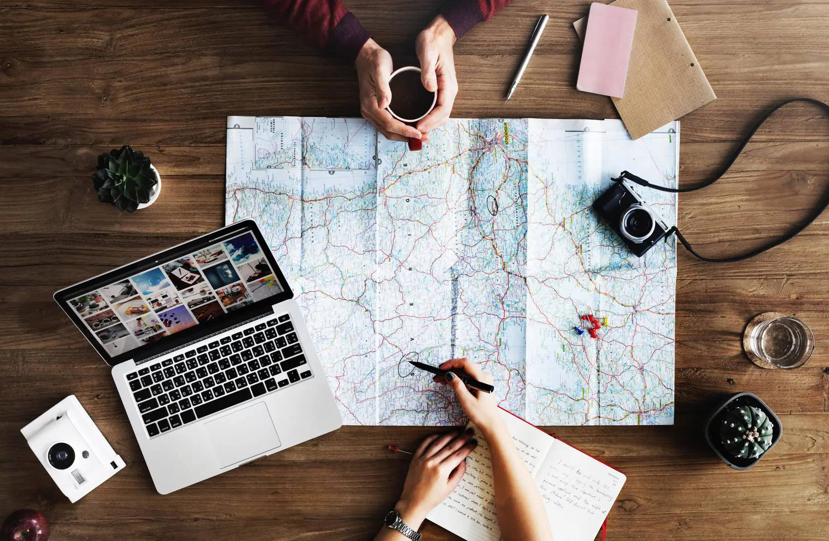 Two people engage in an investigation at a wooden table, with a large map spread out. One person holds a coffee cup, while another writes in a notebook. A laptop, camera, plant, and stationery complete the scene.