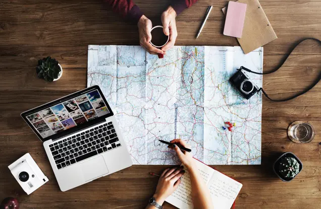 Two people engage in an investigation at a wooden table, with a large map spread out. One person holds a coffee cup, while another writes in a notebook. A laptop, camera, plant, and stationery complete the scene.