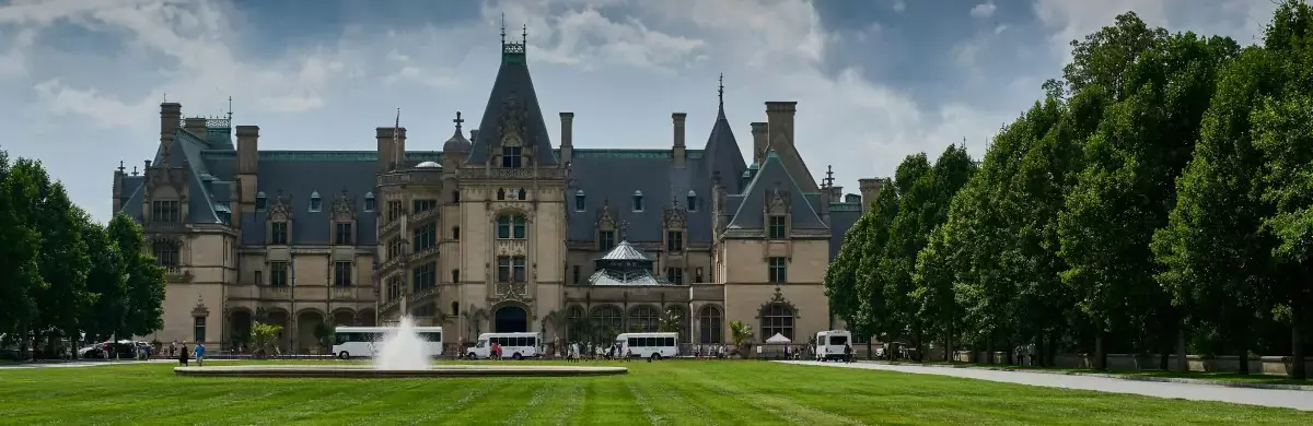 A grand, historic mansion with towers and intricate architecture is surrounded by lush green lawns and trees. An ongoing investigation adds intrigue as a fountain graces the foreground, while several tour buses are parked nearby under a partly cloudy sky.