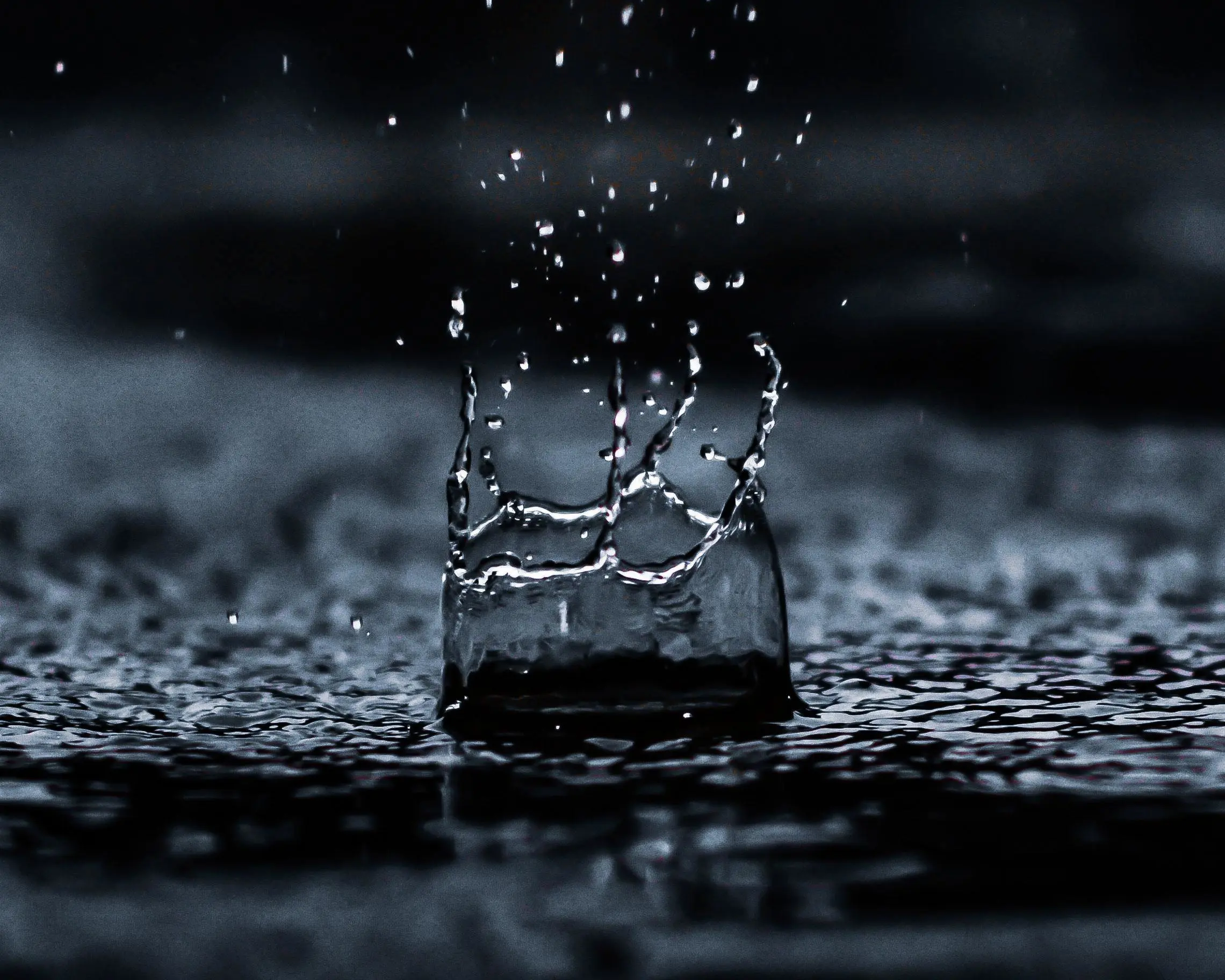 A close-up of a water drop splashing as it hits a dark surface, akin to an inspection in nature, creates a crown-like shape with droplets scattering upwards. The background is softly blurred, emphasizing the motion and impact of this natural assessment.