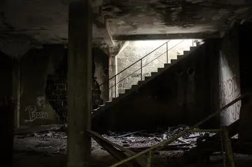 Dimly lit, the abandoned interior hosts a concrete staircase leading upward. The walls are cloaked in graffiti, and debris mingles with patches of mold on the floor. Sunlight peeks through a small opening, casting shadows across the dark space ripe for investigation.